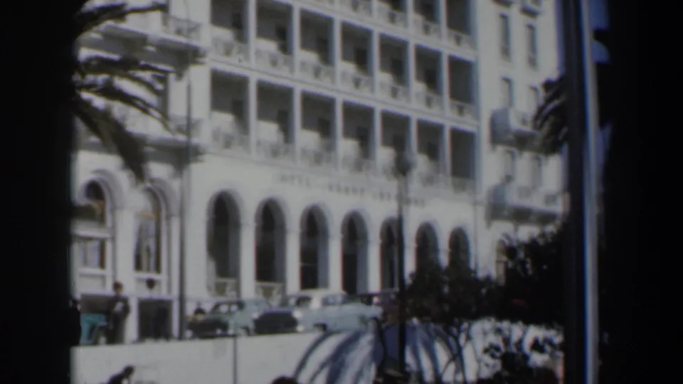 1960 Israel street view of the Hotel Grand Bretagne exterior, with pedestrians, parked cars, arched windows, balconies, and palm fronds.