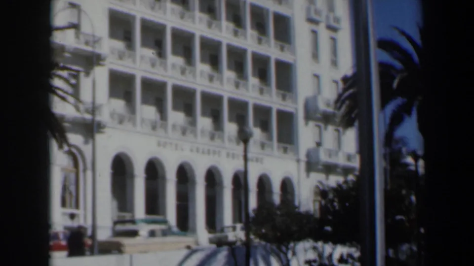 1960 Israel frame with a partially obstructed view of the Hotel Grand Bretagne, showing arches, balconies, cars, palm tree, and pedestrians.