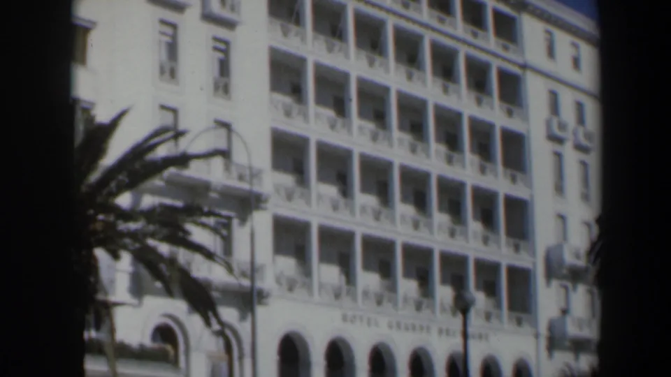 1960 Israel home-movie frame of the Hotel Grand Bretagne facade, with rows of balconies, arched windows, palm fronds, and dark foreground obstruction.