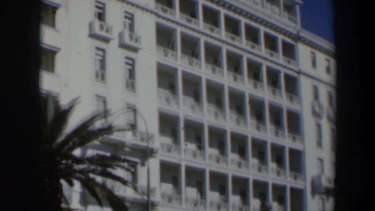 White hotel facade with balconies, windows, palm fronds, and dark film edge.