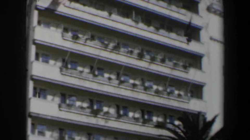 1960 Israel 8mm frame of the Hotel Grande Bretagne exterior, with balconies, awnings, windows, and palm fronds at the edge.
