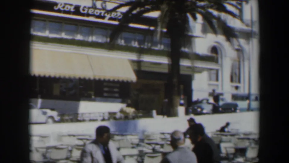 1960 Israel street-level hotel view with Roi Georges signage, parked cars, awnings, palm tree, and cafe patrons.