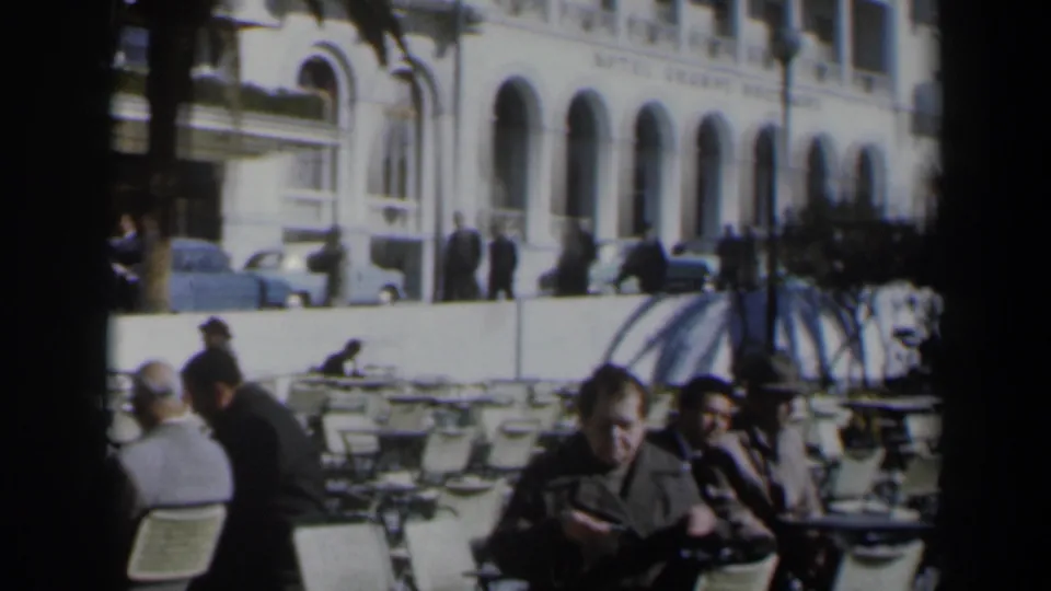 1960 Israel outdoor cafe scene with seated adults, pedestrians, parked cars, and arched hotel frontage behind them.