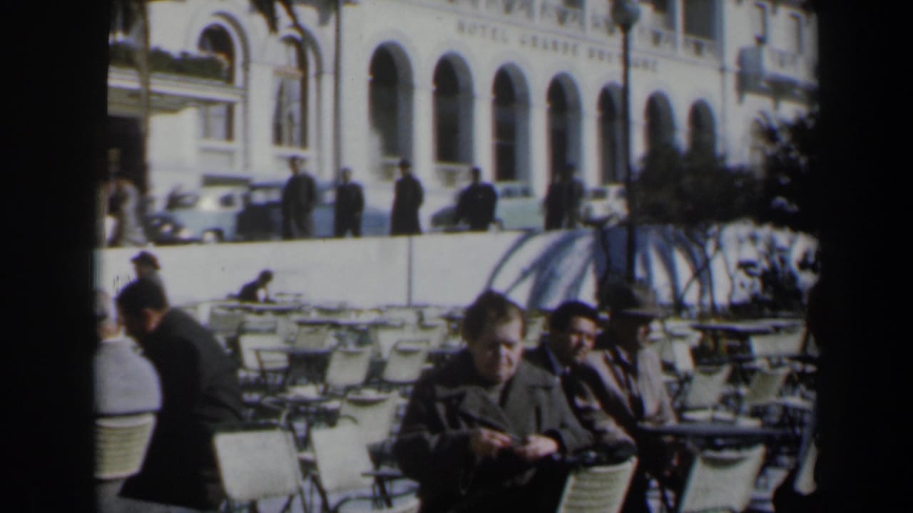Outdoor café with seated men, white metal chairs, pedestrians, cars, and an arched building behind them.