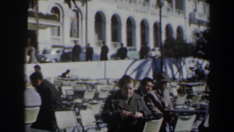 1960 Israel outdoor café scene with seated men, rows of white chairs, pedestrians, vintage cars, and a large arched façade in the background.