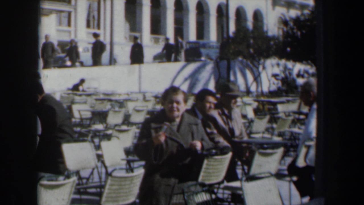 Man in a dark coat gestures near café chairs while other seated patrons and pedestrians appear behind him.