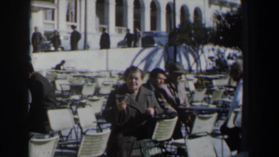 1960 Israel café view with a man gesturing in the foreground, seated patrons, white chairs, pedestrians, cars, and an arched building.