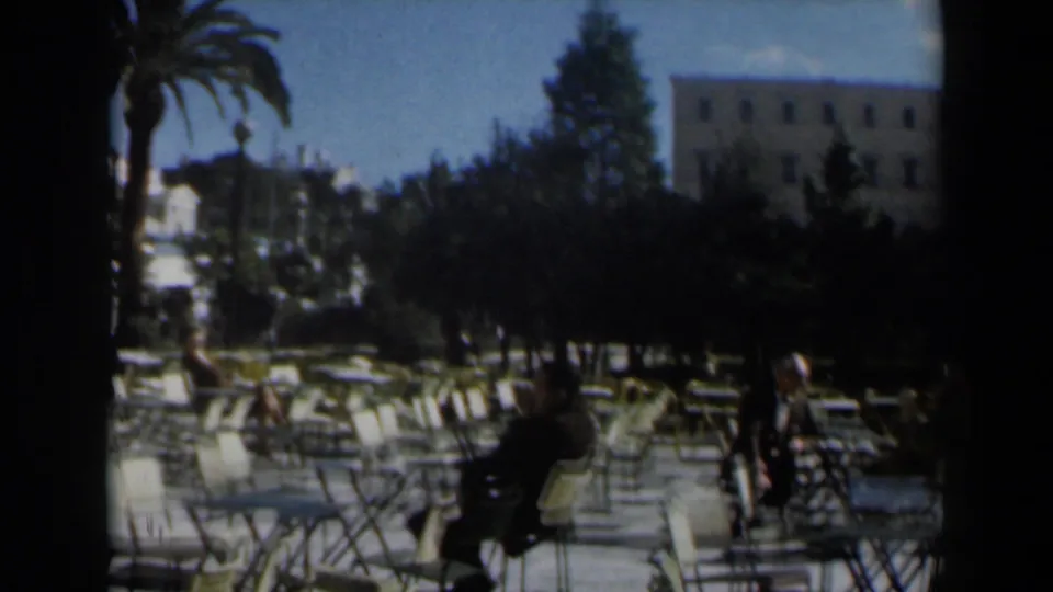 1960 Israel wide café terrace with rows of white chairs and tables, trees, a palm, blue sky, and a few seated figures in shadow.