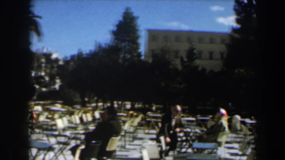 1960 Israel café terrace with scattered seated people, rows of empty white chairs, trees, blue sky, and a large building behind.