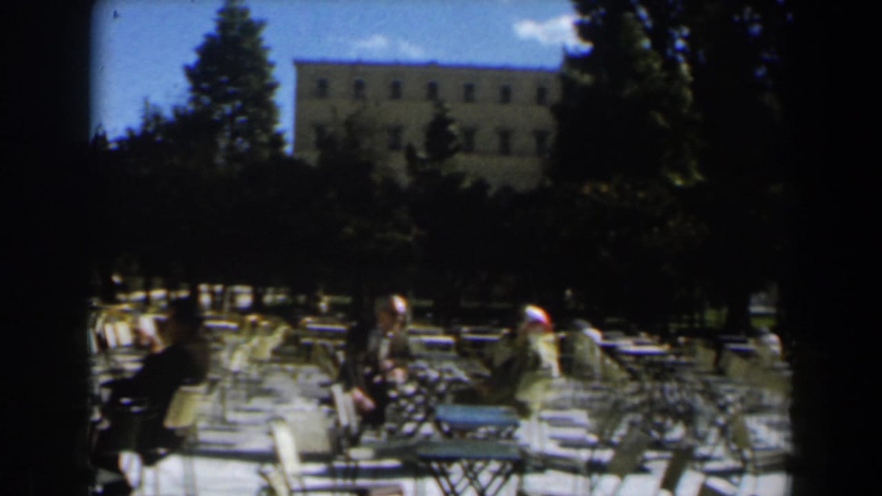 1960 Israel home-movie frame showing a shaded outdoor cafe area with metal seating, adults, trees, and a large pale building in the background.