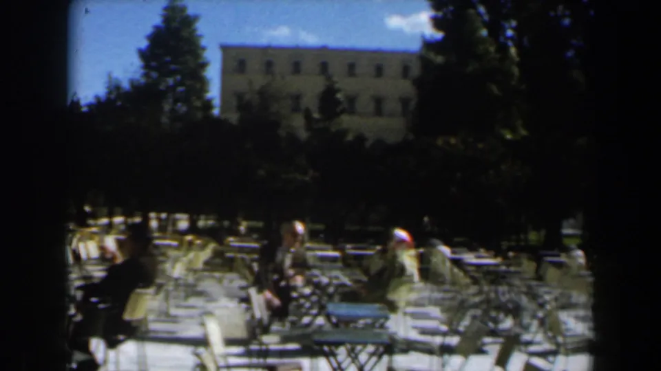 1960 Israel home-movie frame showing a shaded outdoor cafe area with metal seating, adults, trees, and a large pale building in the background.