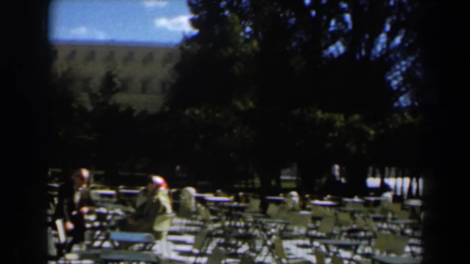 1960 Israel park cafe scene with adults seated near metal tables and chairs, shaded trees, and a large background building.