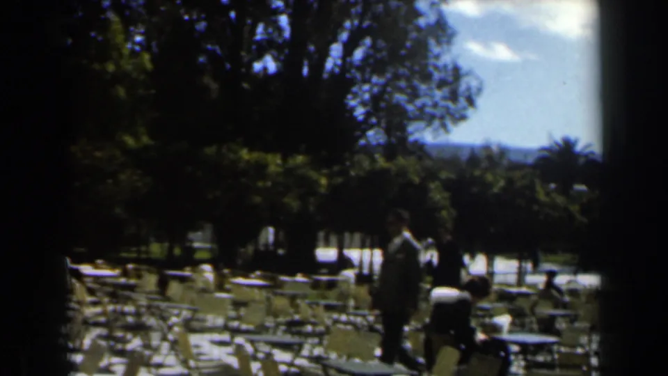 1960 Israel home-movie view across a park cafe area, with empty chairs, tables, trees, and an adult standing near the walkway.
