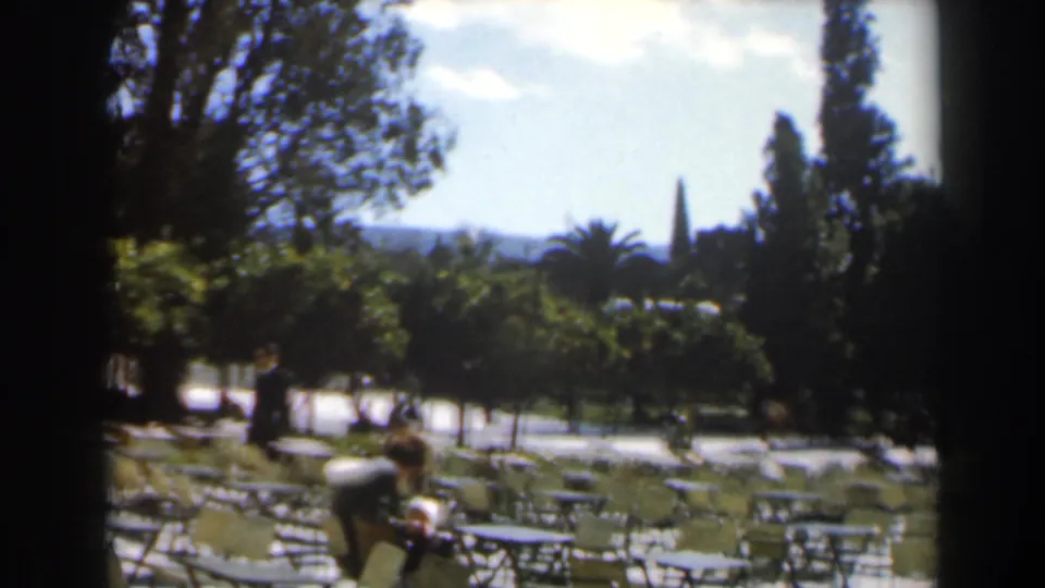 1960 Israel park cafe frame showing adults near metal tables, shaded trees, garden paths, and a distant skyline or hillside.