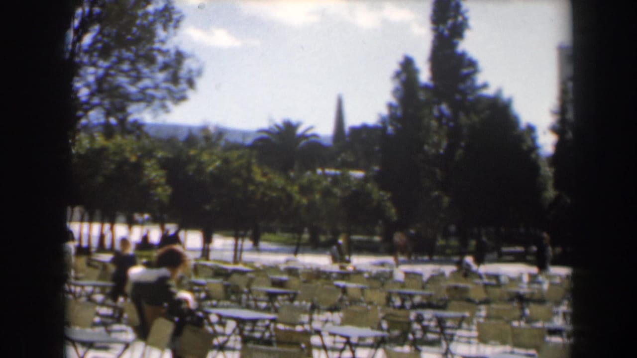 1960s Israel home-movie frame showing an outdoor park or cafe seating area with trees, empty chairs, and a few distant people.