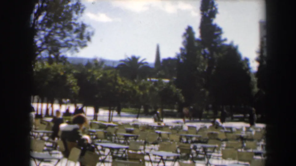 1960s Israel home-movie frame showing an outdoor park or cafe seating area with trees, empty chairs, and a few distant people.