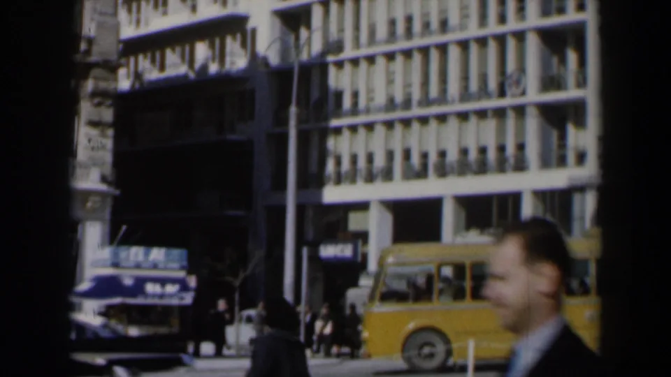 1960s Israel street scene with a white building, yellow bus, dark vehicle, pedestrians, and a blurred man in the foreground.