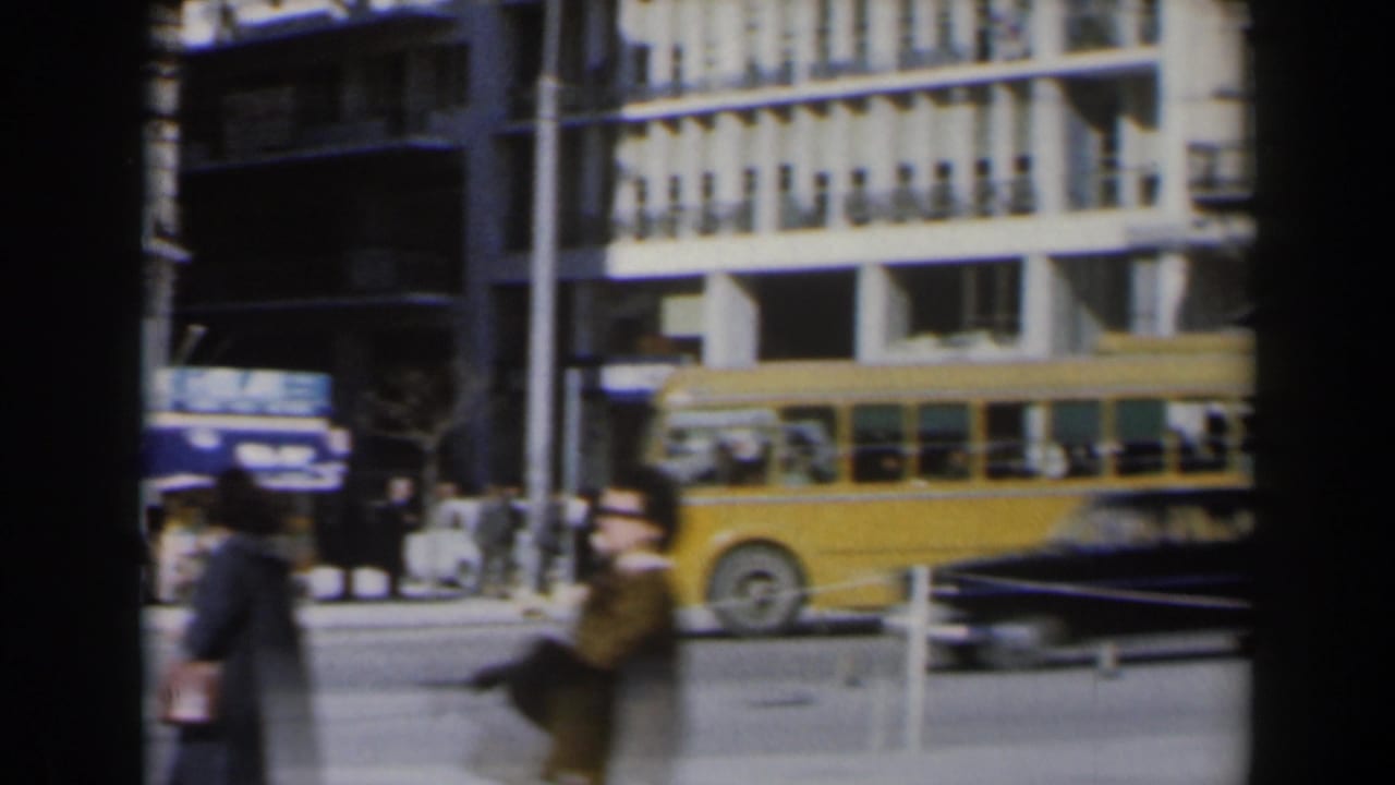 Pedestrians and a yellow bus pass a commercial street seen through dark film-edge bands.