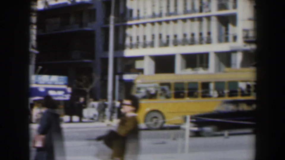 1960 archival home-movie frame of an Israeli city street with pedestrians, a yellow bus, older cars, and commercial buildings.