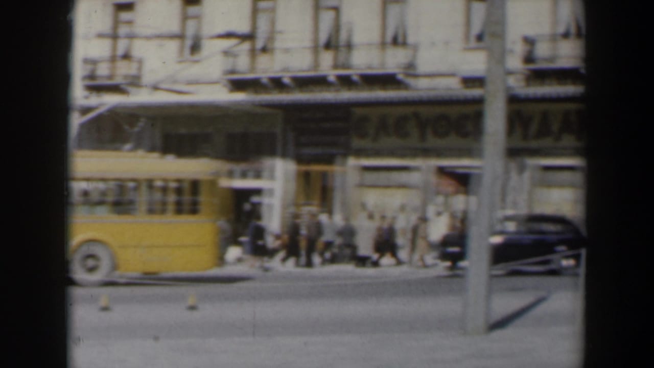 Yellow bus, pedestrians, and cars pass storefronts with Greek-letter signage.