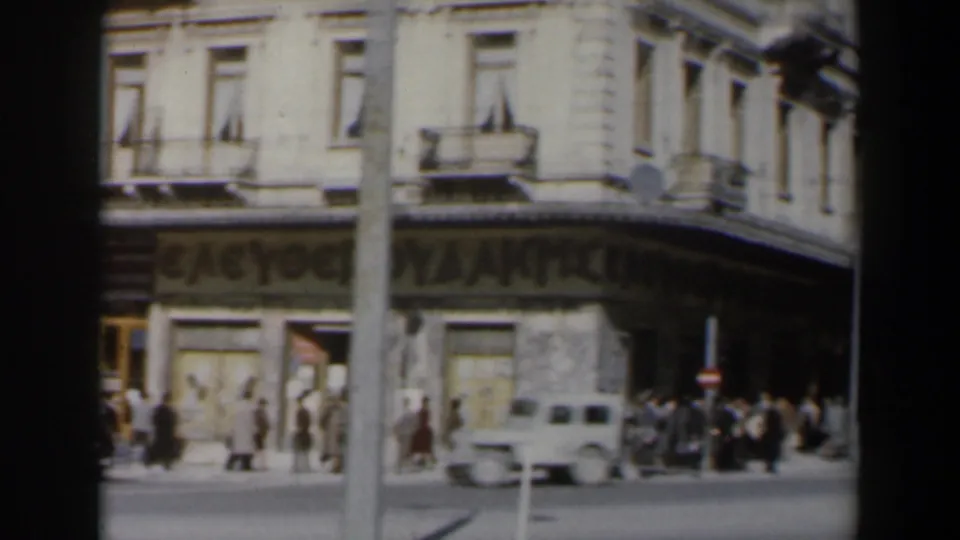 1960 home-movie street view in Israel with pedestrians gathered near a corner shopfront, balconies, signage, and a passing light car.