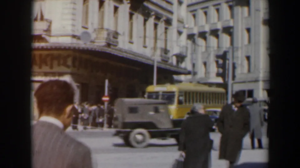 A 1960 Israel city frame shows men in coats near the camera, a yellow bus, a dark utility vehicle, pedestrians, and commercial facades.
