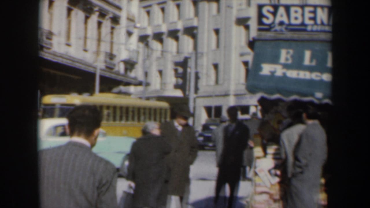 1960s Israel home-movie frame of suited pedestrians on a city sidewalk, with a yellow bus, older cars, and a SABENA sign above the street.
