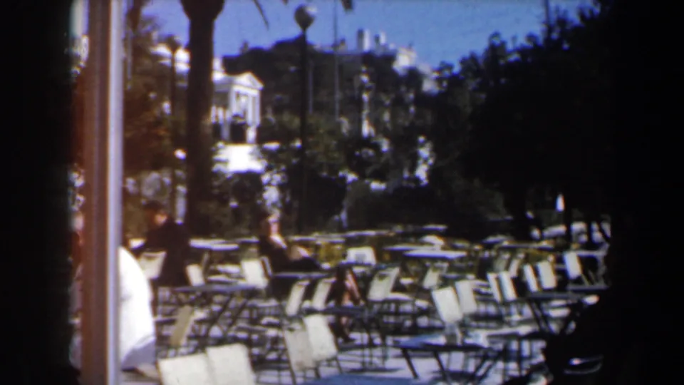 1960s Israel outdoor seating area with many empty cafe chairs, trees, pedestrians, and bright buildings in the background.