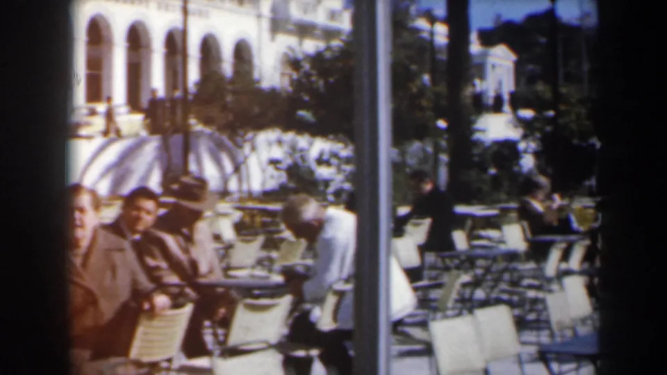 Outdoor cafe scene in Israel with seated people, empty chairs, a striped awning, trees, and an arched building facade in soft 8mm color.