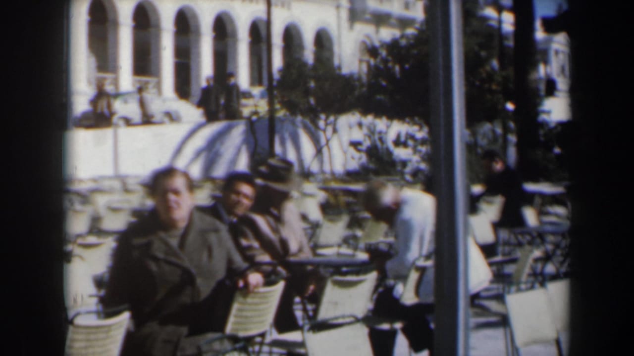 Adults sit at folding café tables and chairs near umbrellas and an arched building.