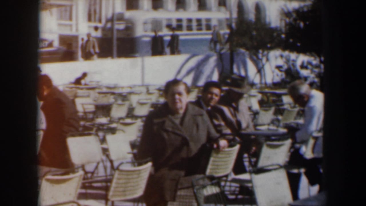 Two adults face the camera while seated among café tables and chairs outdoors.