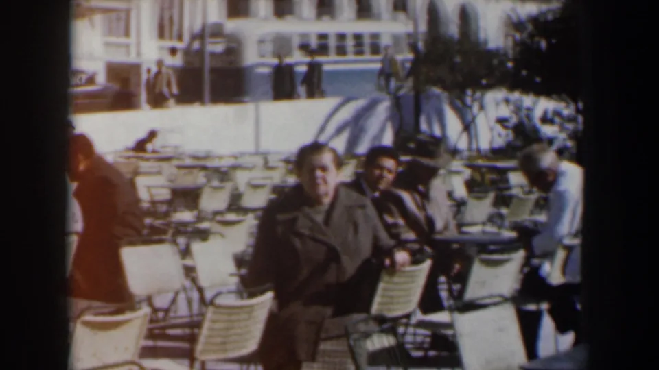 Two adults sit at outdoor café furniture in Israel, with pedestrians, umbrellas, and a large building visible in the 1960 home-movie frame.