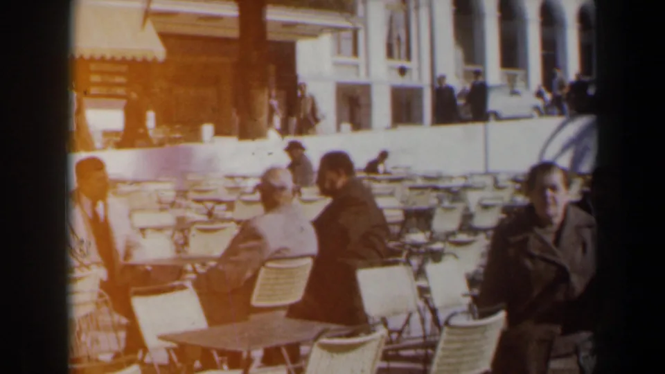A 1960 Israel frame shows adults gathered around folding café tables, with warm film flare, umbrellas, and a street-side building in view.