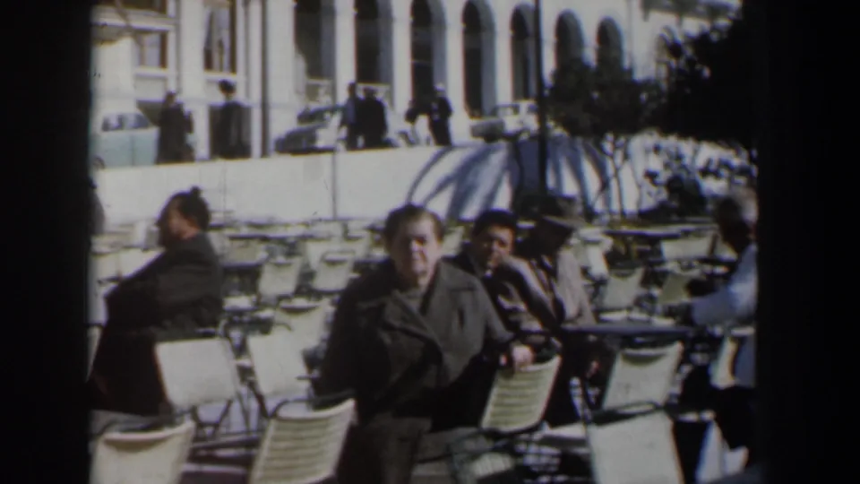 Adults remain seated in an outdoor café setting in 1960 Israel, with folding chairs, striped umbrellas, arched windows, and street activity behind them.
