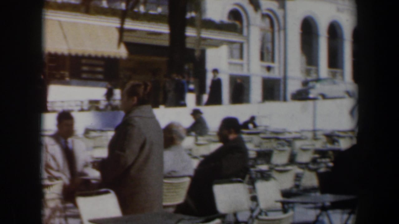 Outdoor cafe terrace with seated patrons, wicker chairs, and a woman in a dark coat near the foreground.