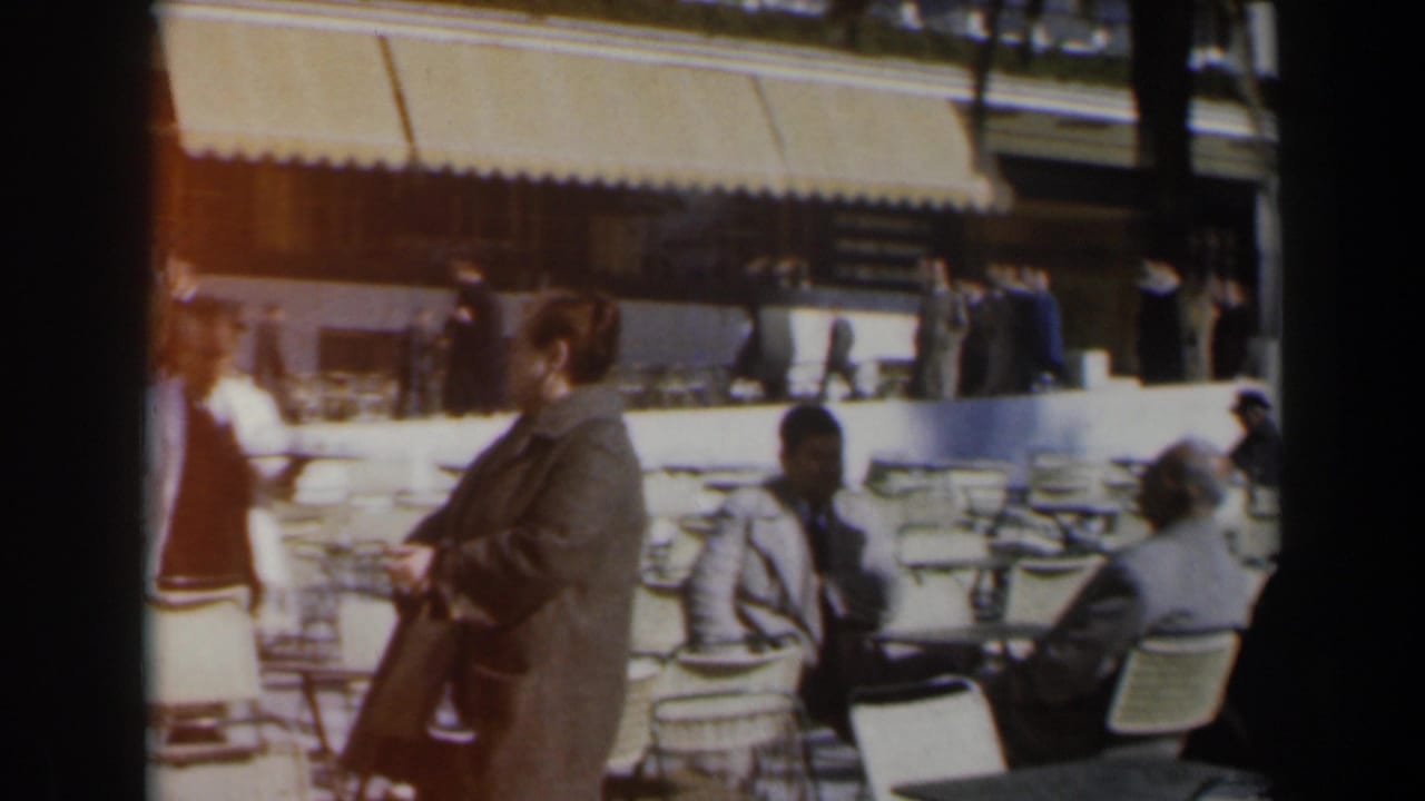 Woman in a dark coat turns near outdoor cafe tables as people sit and walk behind her.