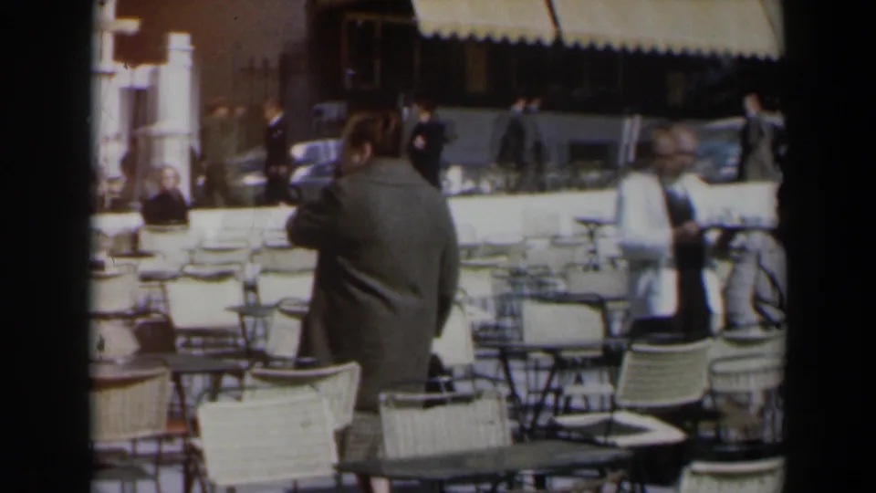 1960 Israel home-movie frame from behind a woman leaving an outdoor cafe, with chairs, walkers, and mid-century cars in the background.