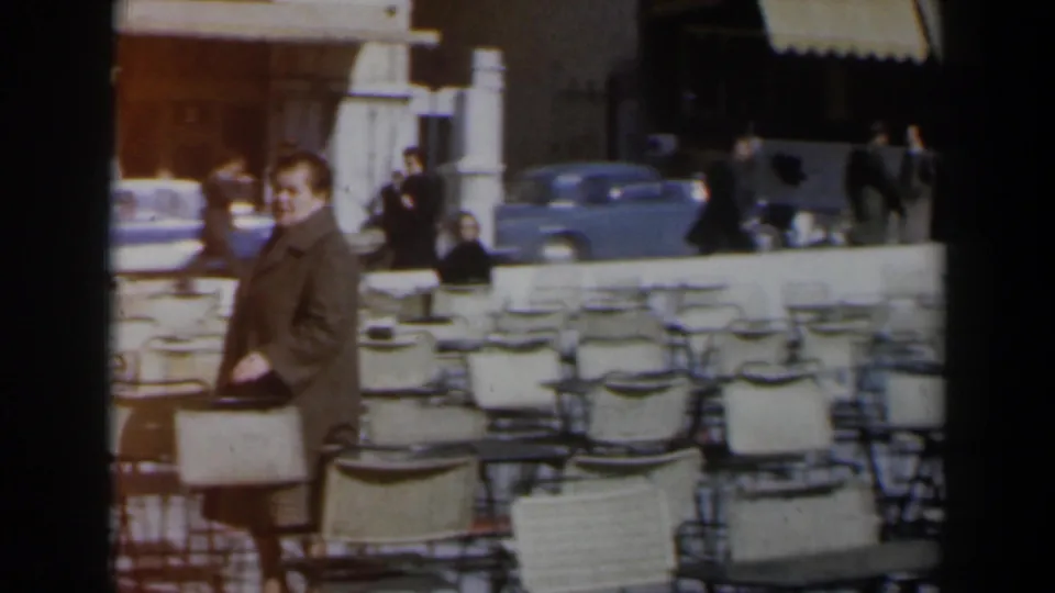 1960 Israel frame showing a woman near an outdoor cafe looking toward the camera, with wicker seating, pedestrians, and parked cars behind her.