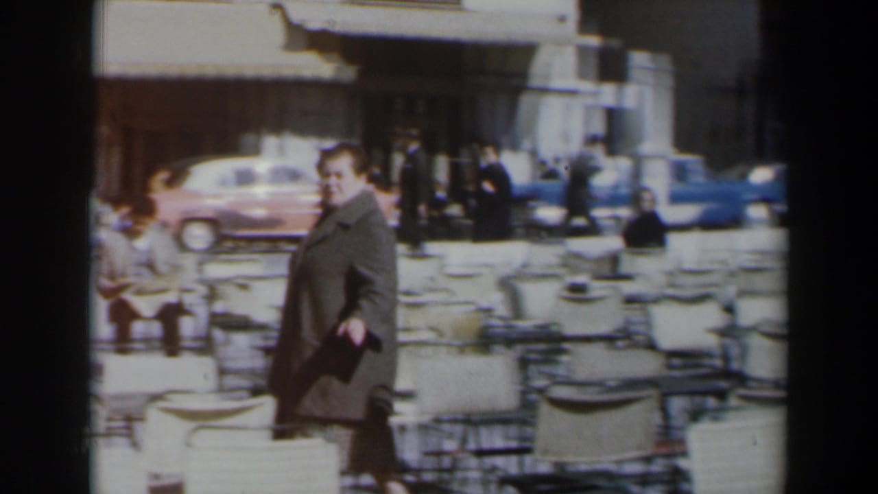 Woman in a grey coat walks past white cafe chairs on a sunny sidewalk.