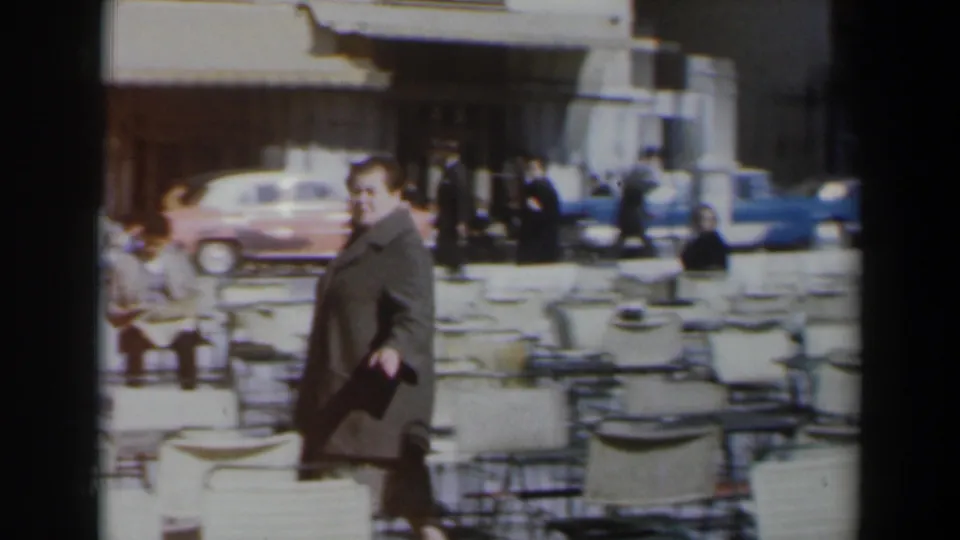 In 1960s Israel, a woman in a grey coat walks through outdoor cafe seating with parked cars and pedestrians behind her.