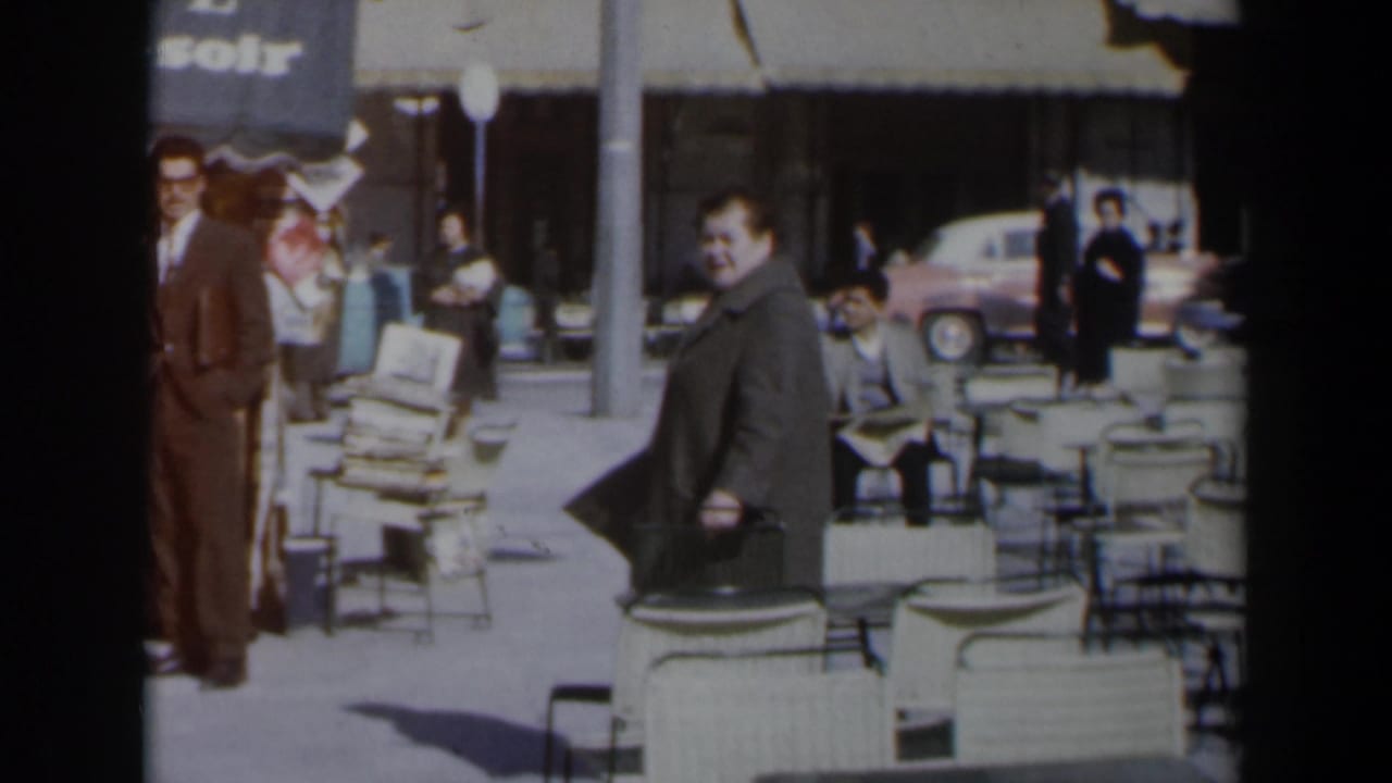 Woman in grey coat stands or walks near cafe chairs as people gather nearby.