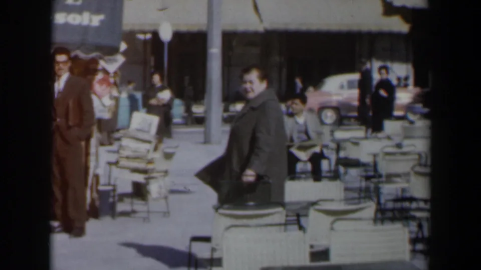 The woman in a grey coat moves near white outdoor chairs while men in suits and other pedestrians appear on the sidewalk in 1960s Israel.