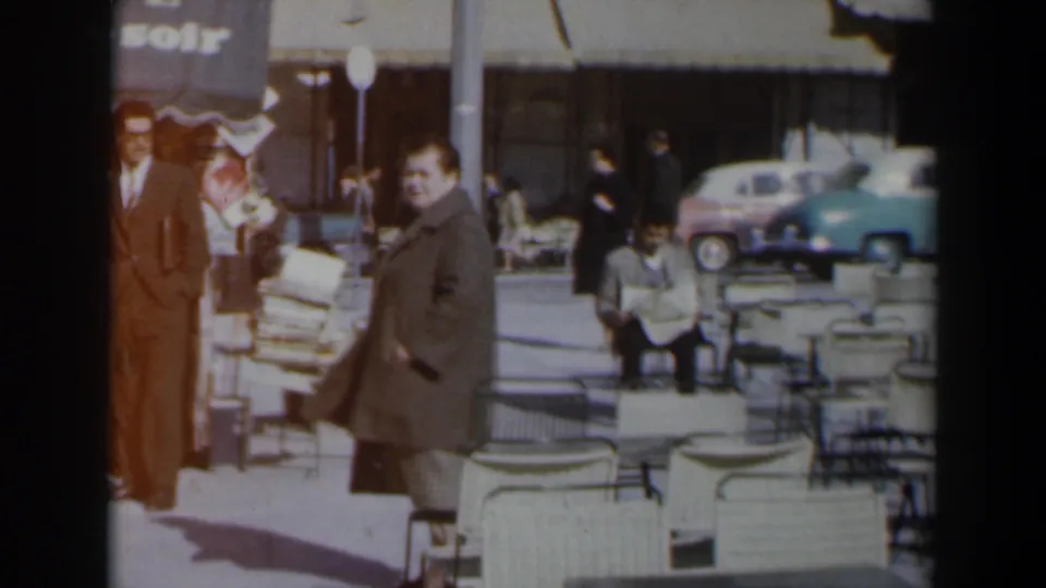 In a 1960s Israel street scene, a woman in a grey coat walks past a man in a brown suit near white cafe furniture and parked cars.