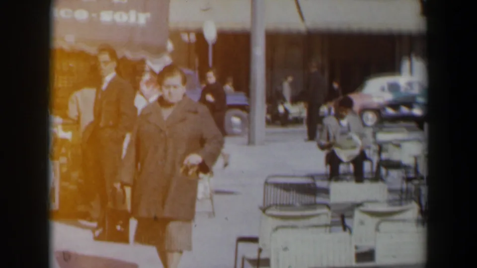 A woman in a grey coat walks toward the camera in 1960s Israel, with men, white cafe chairs, parked cars, and a partial storefront sign behind her.