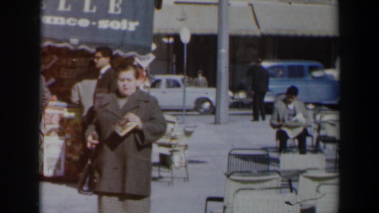 Woman in a brown coat walks on a sunny sidewalk holding a book and handbag.