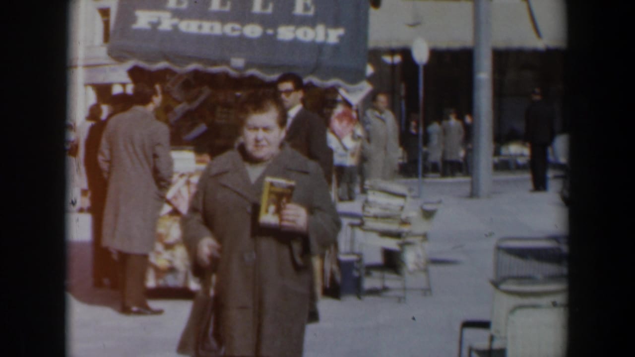 Woman holding a book passes a sidewalk kiosk with pedestrians and cars behind her.