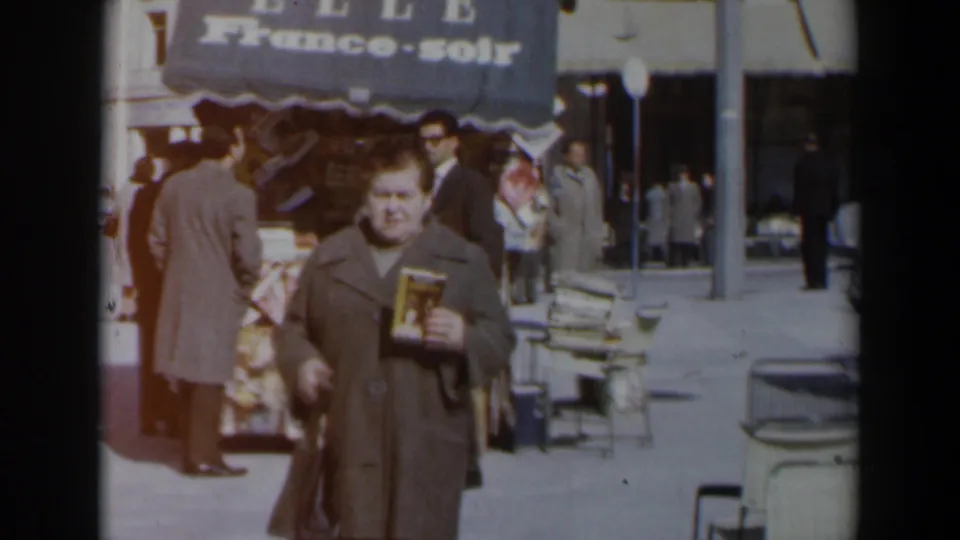 A woman carrying a book and handbag walks along a sunlit Israeli street, with pedestrians, parked cars, and a France-soir awning behind her.
