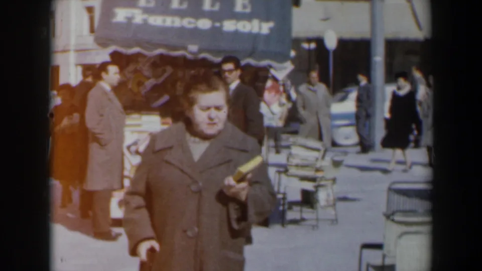 The home-movie frame shows the woman closer to camera, carrying a book beside cafe chairs and a busy sidewalk in 1960s Israel.