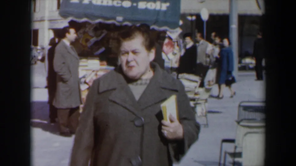 A woman in a brown coat nears the camera with a book tucked in her arm while pedestrians pass behind a France-soir awning.