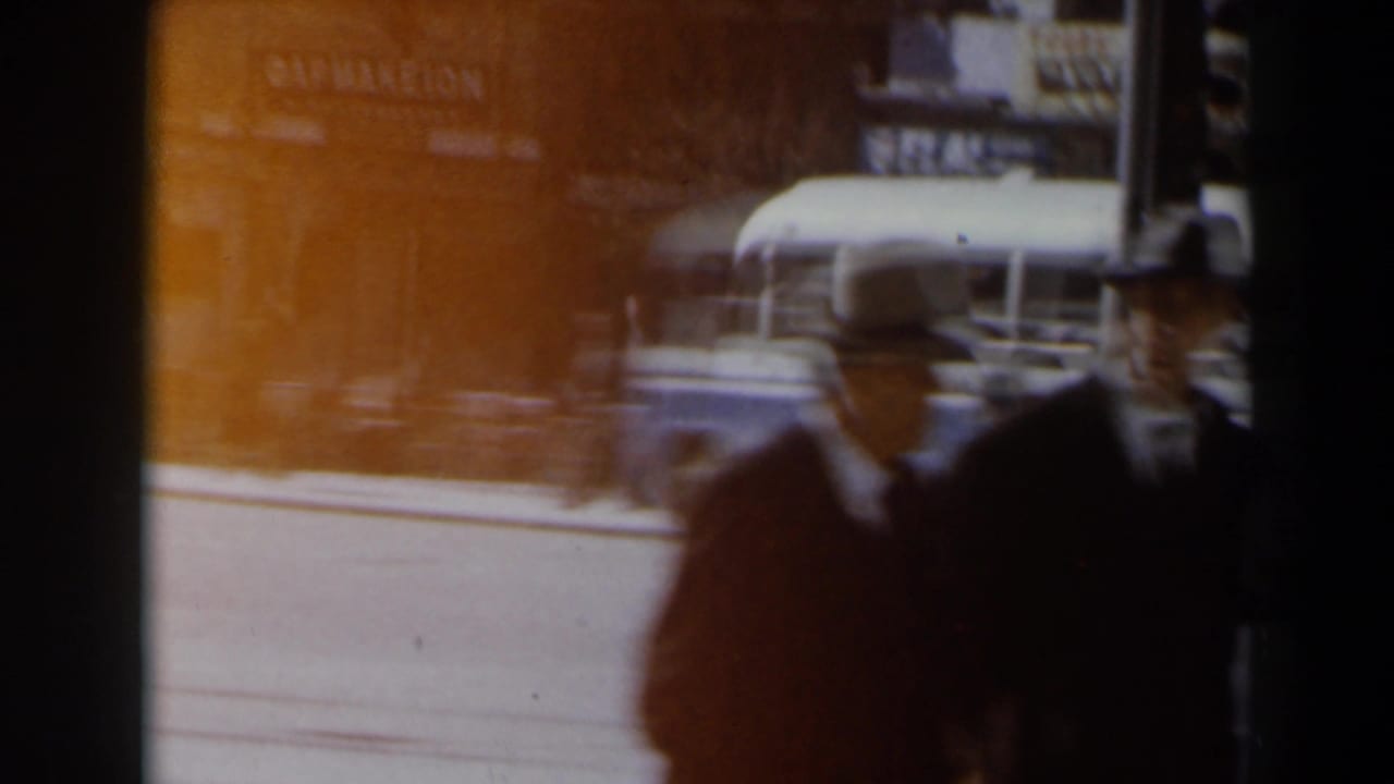 1960s Israel street view with pedestrians in dark clothing, a bright roadway, and a shop awning behind them.